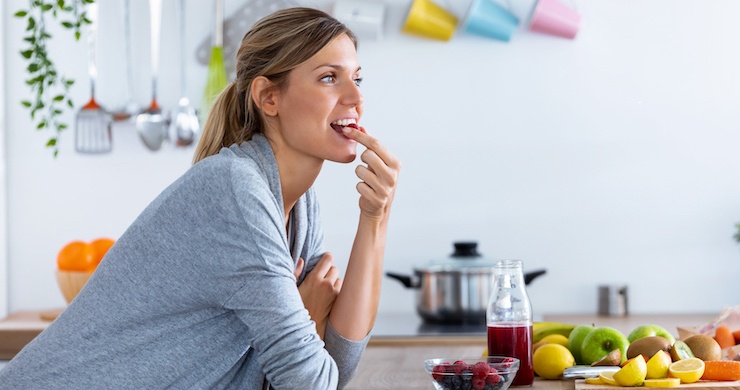 Pretty young woman eating red berries while sitting in the kitchen at home.